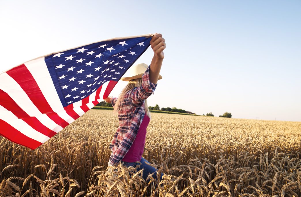 Attractive blonde female walking through wheat field and holding USA waving flag. Powerful economy and agriculture of the United States of America.