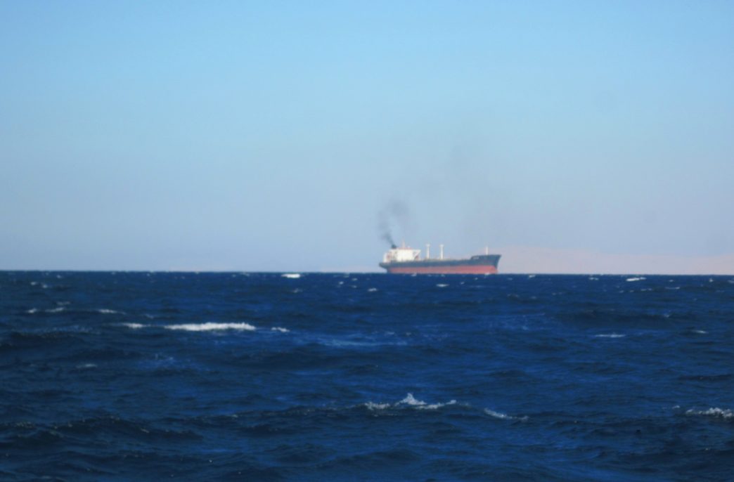 Blue sea surface. A black and red-striped cargo ship floats on the horizon. Black smoke is pouring out of its chimney. A yellow mountain can be seen behind the ship